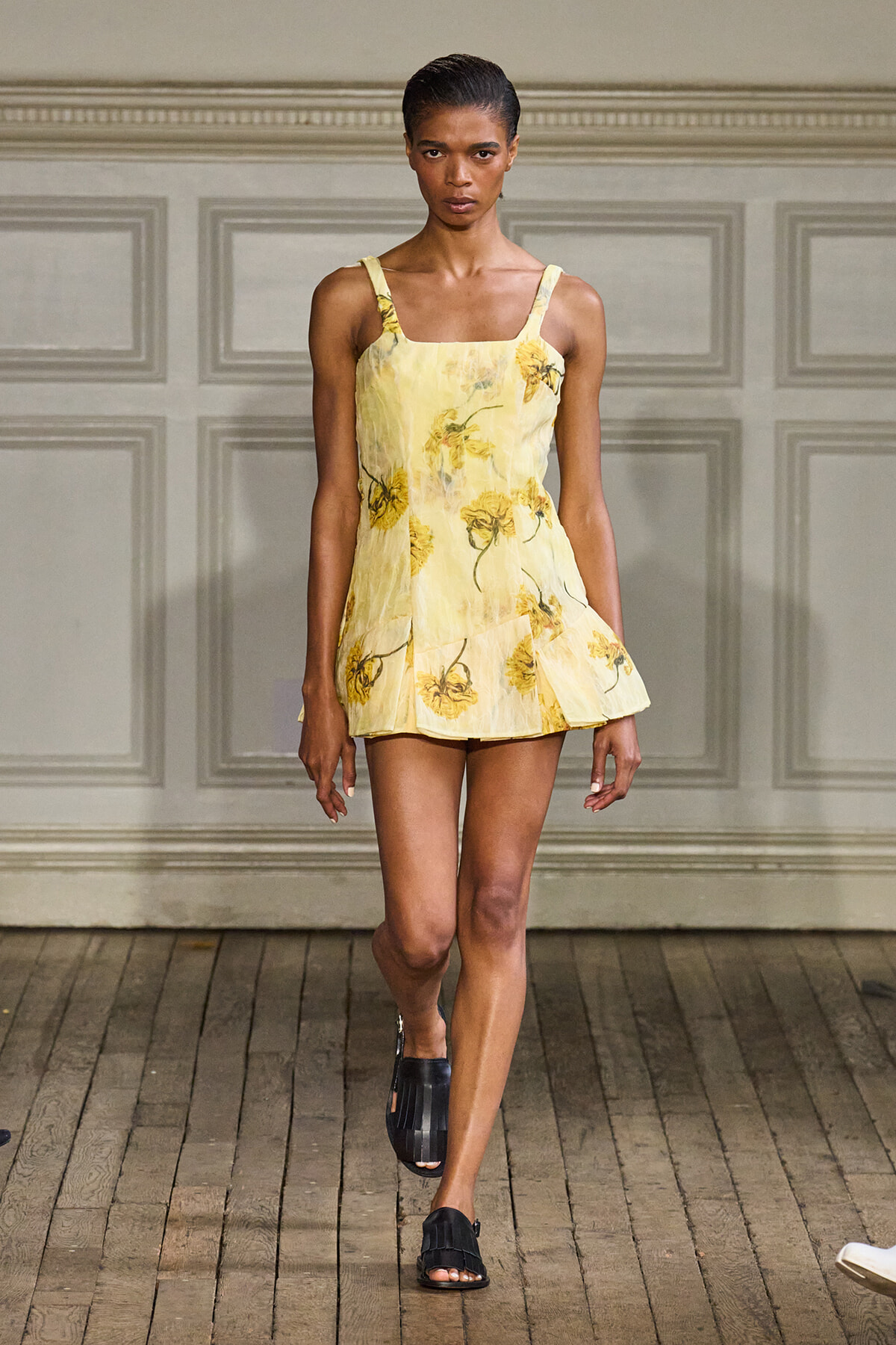 Female model walking a fashion runway in a pale yellow floral sundress with thin straps and black sandals.
