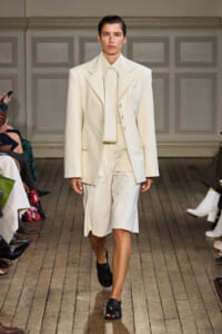 Model walks the runway in an ivory tailored blazer and matching knee-length shorts, paired with a high-collar shirt and black sandals.