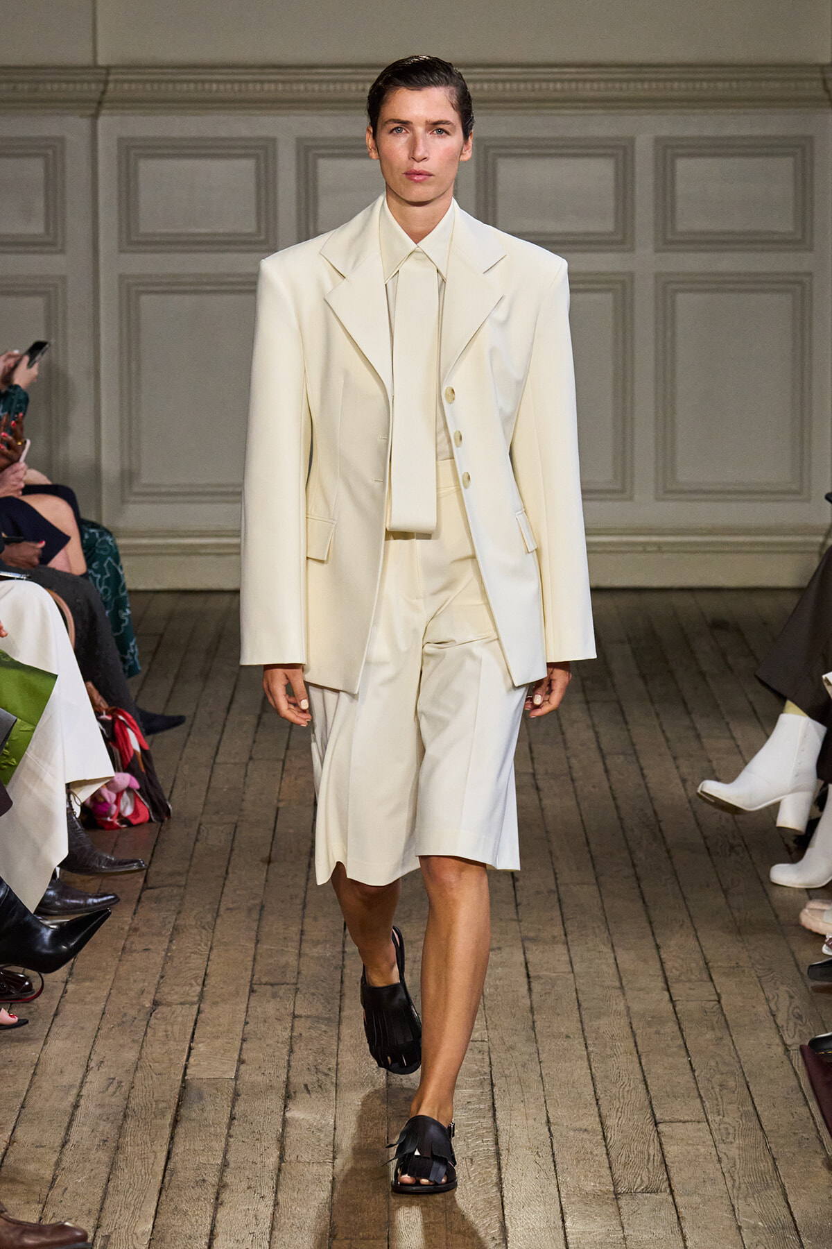 Model walks the runway in an ivory tailored blazer and matching knee-length shorts, paired with a high-collar shirt and black sandals.