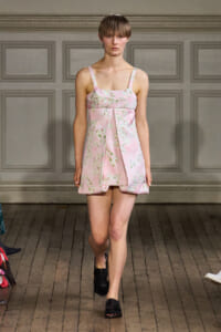 Model walking a runway in a pink floral sleeveless mini dress with a pleated front and square neckline, paired with black slide sandals.