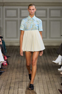 Model walking a runway in a light blue floral blouse tucked into a cream pleated mini skirt with black sandals.