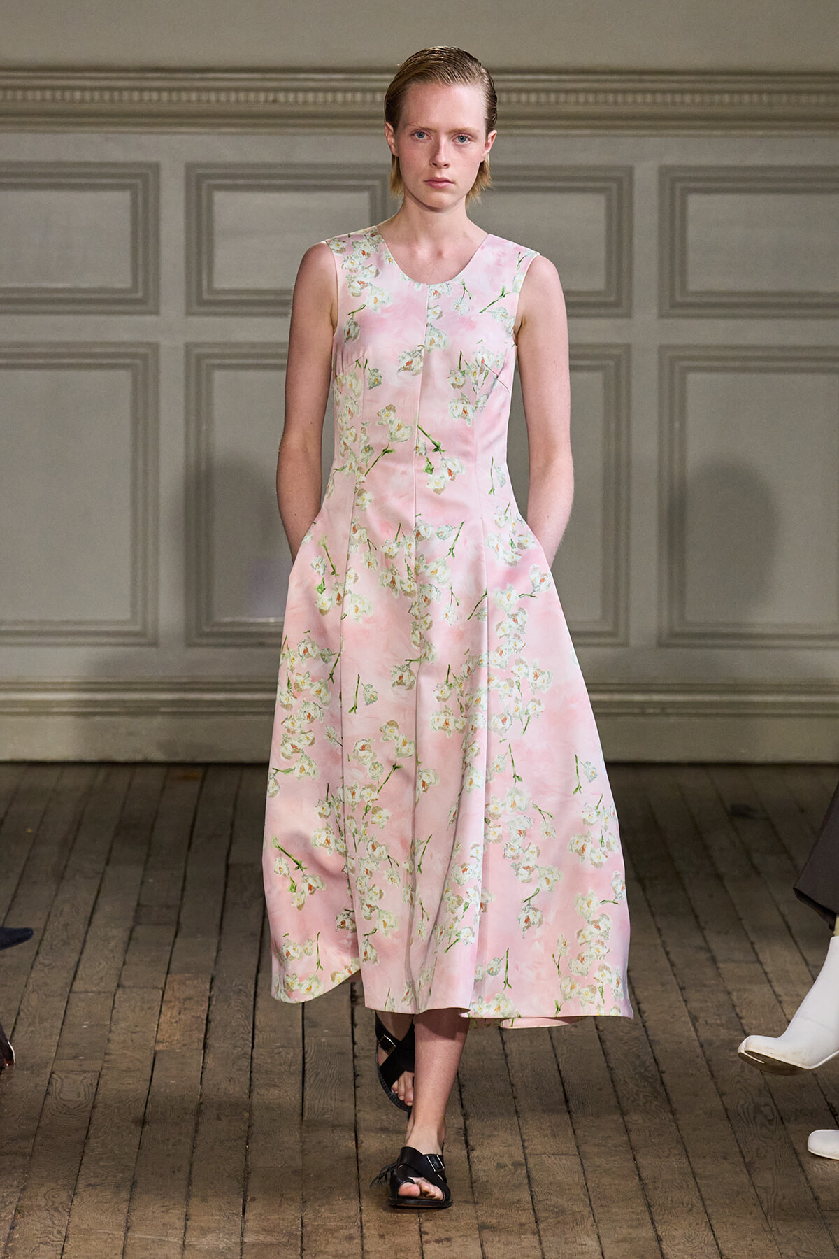 Model walking in a pink sleeveless midi dress with a white floral print on a wooden runway.
