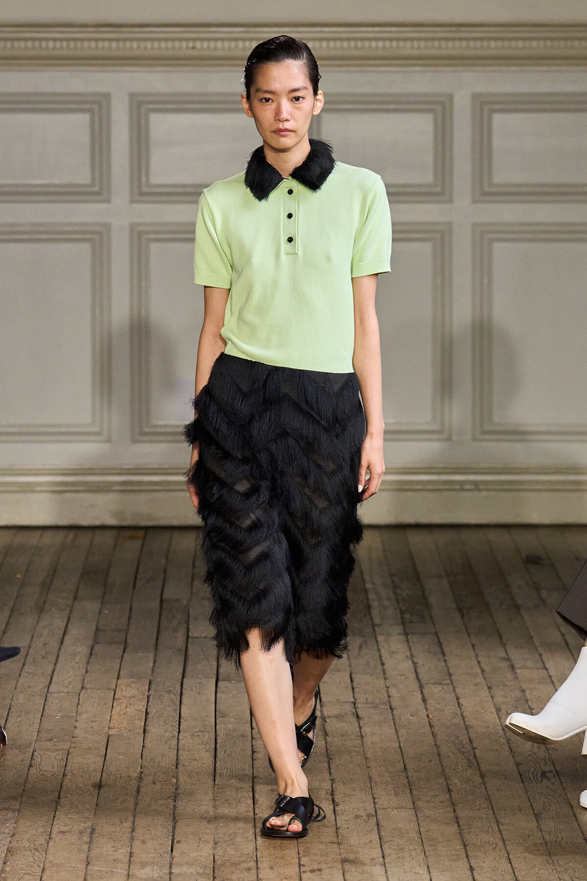 Model walking a runway in a pale green polo top with a black fur-trimmed collar and a layered black feathered skirt, wearing black sandals.