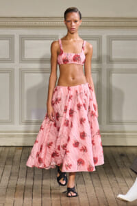 Model on a runway wearing a pink floral two-piece set: cropped top and long full skirt, with black sandals.