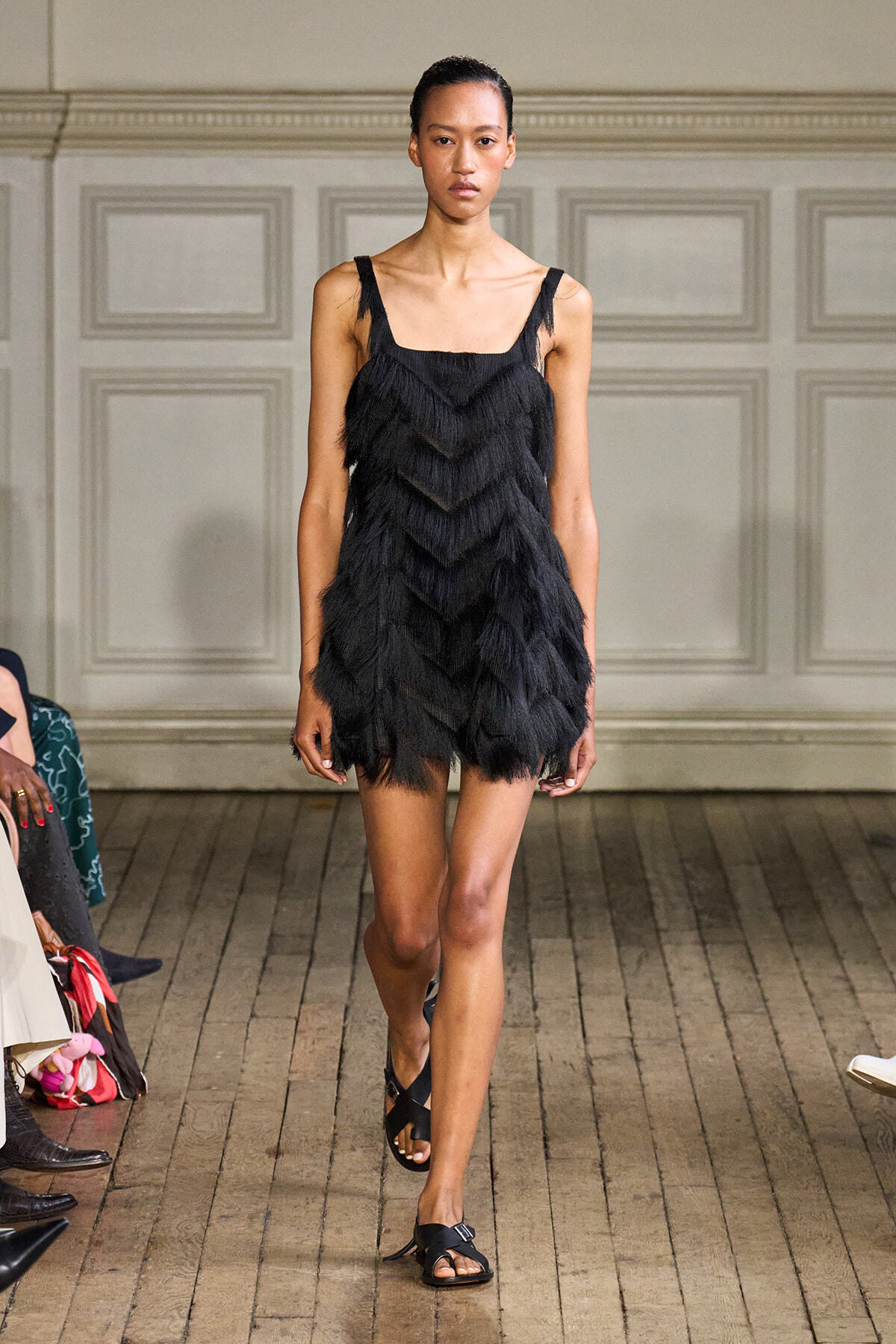Model walks the runway in a black fringe mini dress with thin straps during a fashion show.