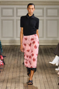 Model walking the runway in a black polo top with a pink floral midi skirt trimmed with black feathers at the hem, wearing black sandals.