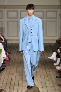 Model on a fashion runway wearing a pale blue oversized suit with broad lapels and a three-button vest, paired with wide-leg trousers.