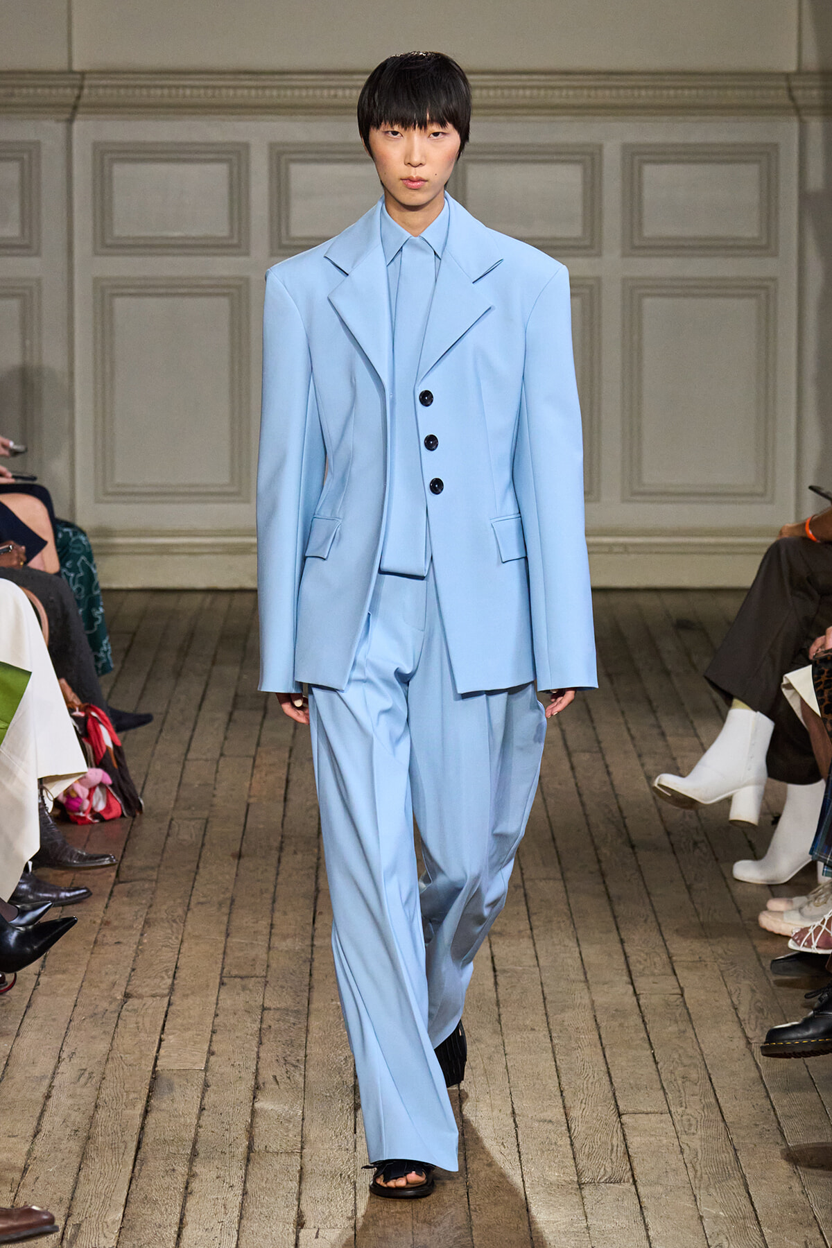 Model on a fashion runway wearing a pale blue oversized suit with broad lapels and a three-button vest, paired with wide-leg trousers.