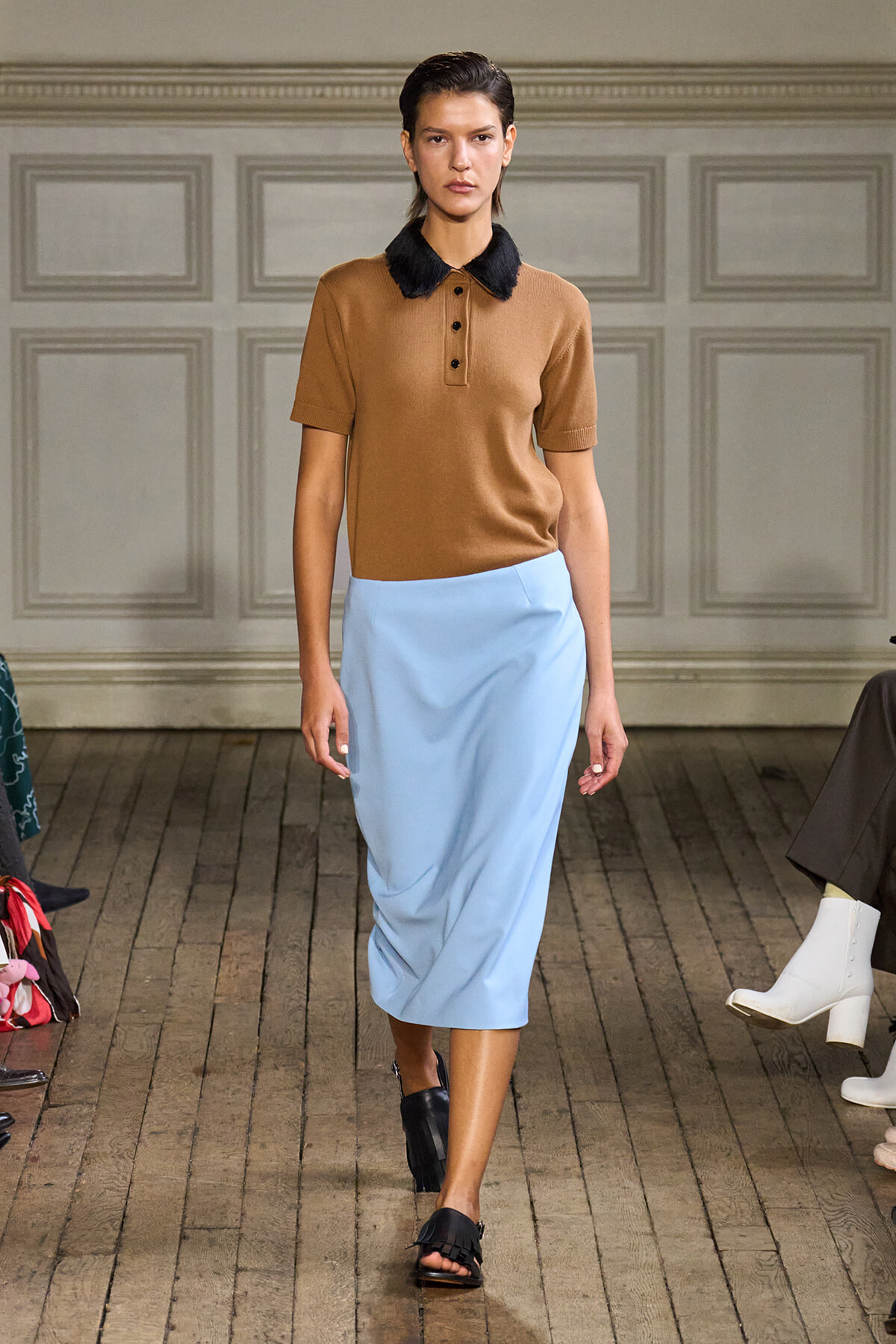 Model wearing a camel polo with a black fur collar and a powder blue midi skirt on a runway runway walk (fashion show).