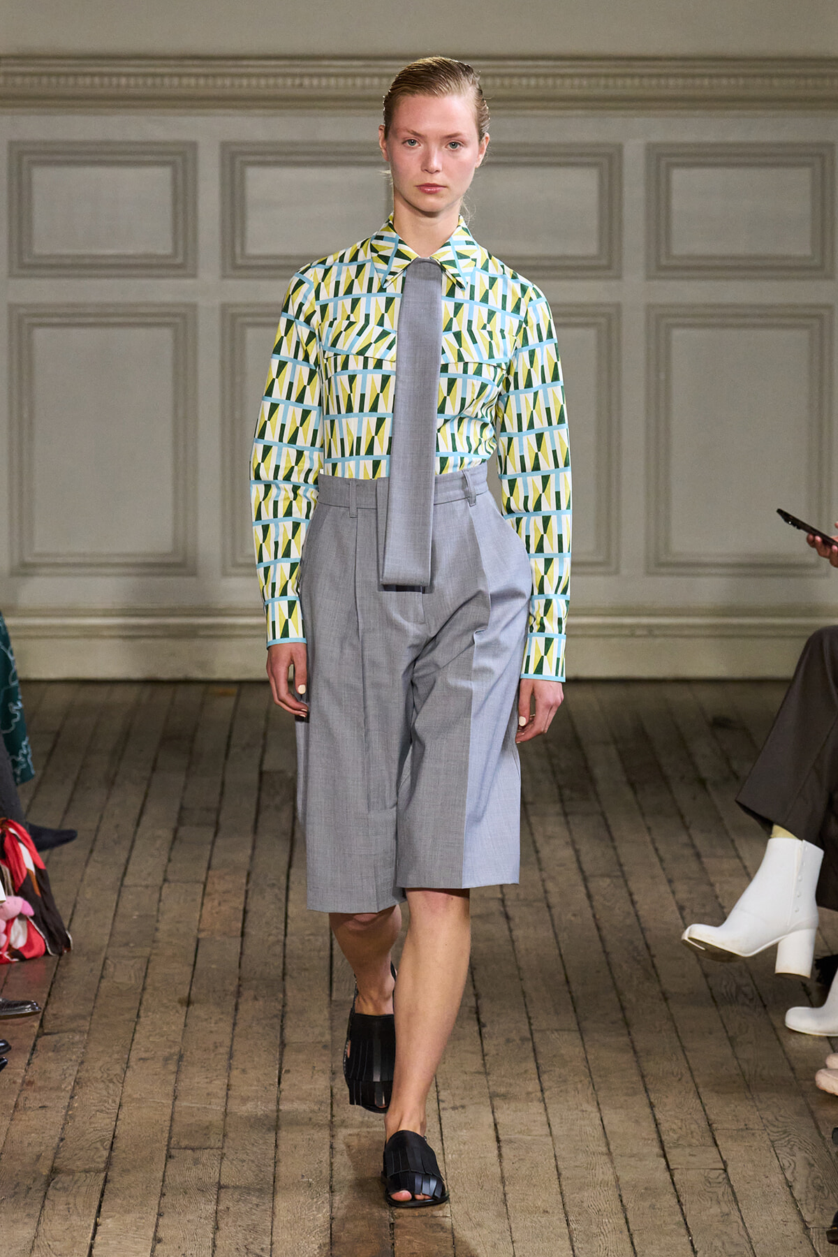 Model on a fashion runway wearing a geometric patterned green, blue, and white shirt with a gray necktie and high-waisted light-gray wide shorts, black sandals.