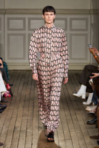 Model walks the runway in a pink and black geometric prints jumpsuit, belt cinching the waist, black sandals.