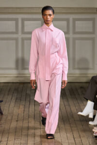 Model walking a fashion runway in a pale pink shirt-style blouse and matching wide-leg pants with a tie belt.