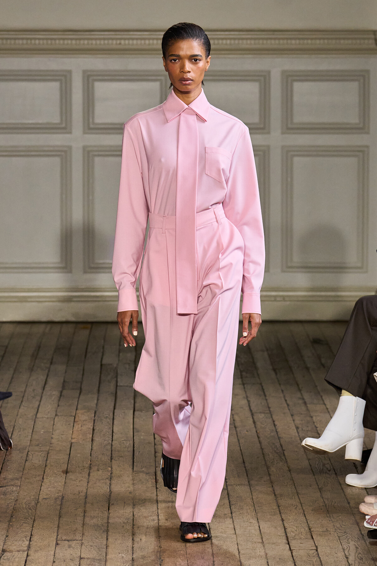 Model walking a fashion runway in a pale pink shirt-style blouse and matching wide-leg pants with a tie belt.
