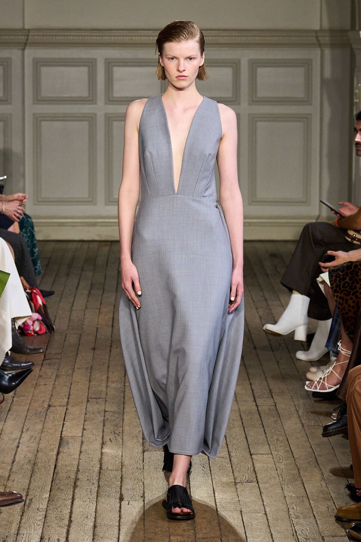 Model walking a fashion runway in a light gray sleeveless deep-V gown with a draped, wide-skirt silhouette.