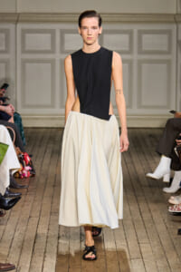 Model walks the runway in a black sleeveless top with an asymmetric cut paired with a voluminous white midi skirt.