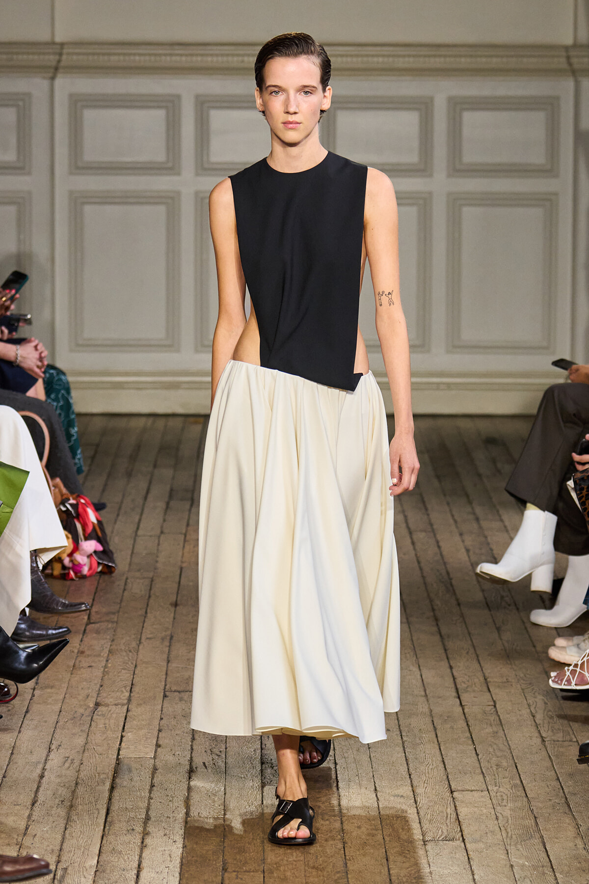 Model walks the runway in a black sleeveless top with an asymmetric cut paired with a voluminous white midi skirt.
