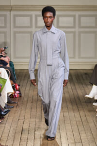 Model walking the runway in a light gray jumpsuit with a wide collar and matching tie, black sandals visible underneath.