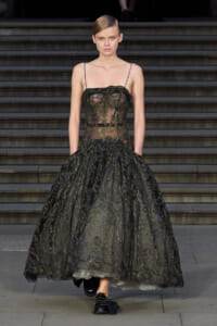 Model walking down a runway in a black lace ball gown with gold embroidery and thin spaghetti straps on a staircase backdrop.