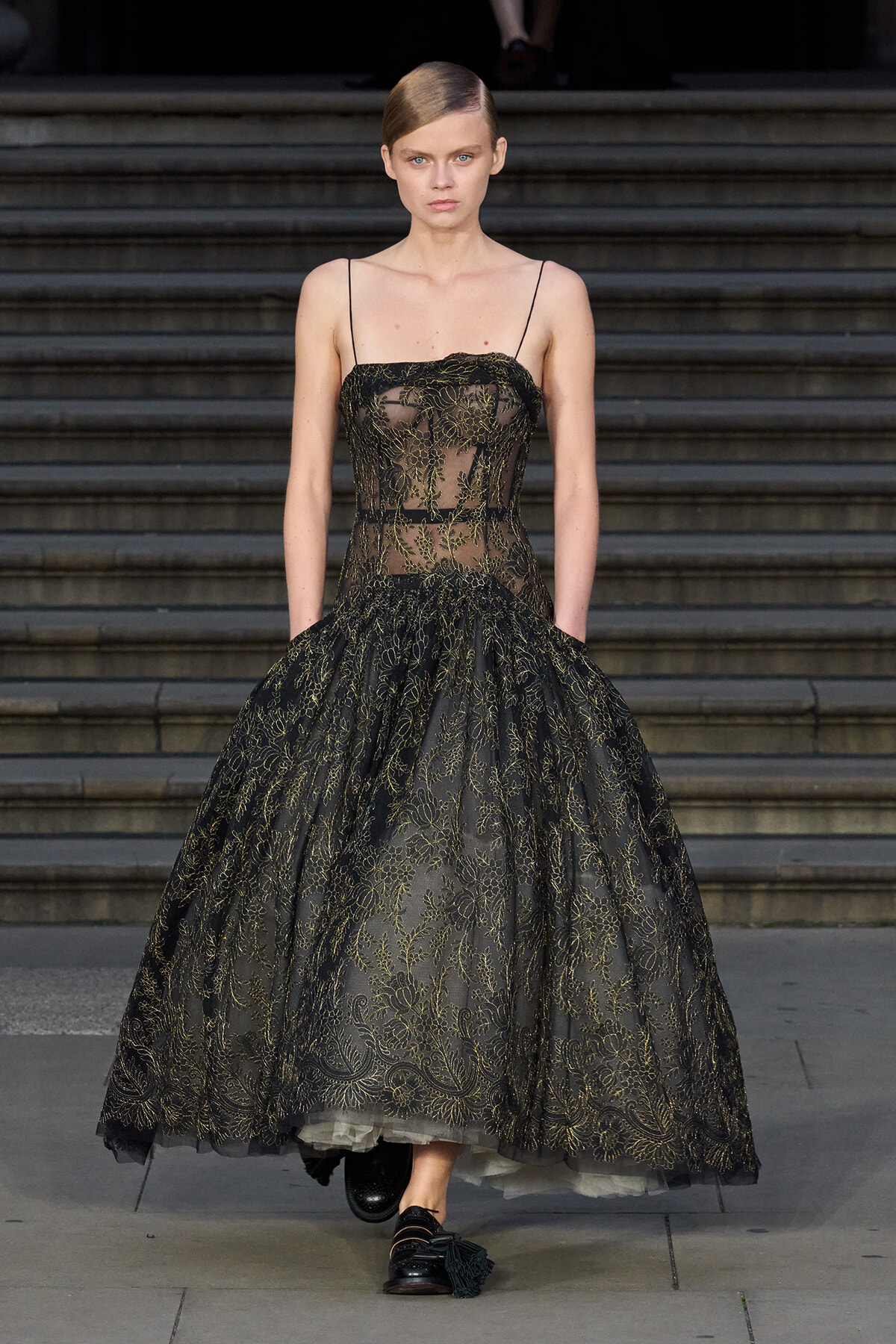 Model walking down a runway in a black lace ball gown with gold embroidery and thin spaghetti straps on a staircase backdrop.