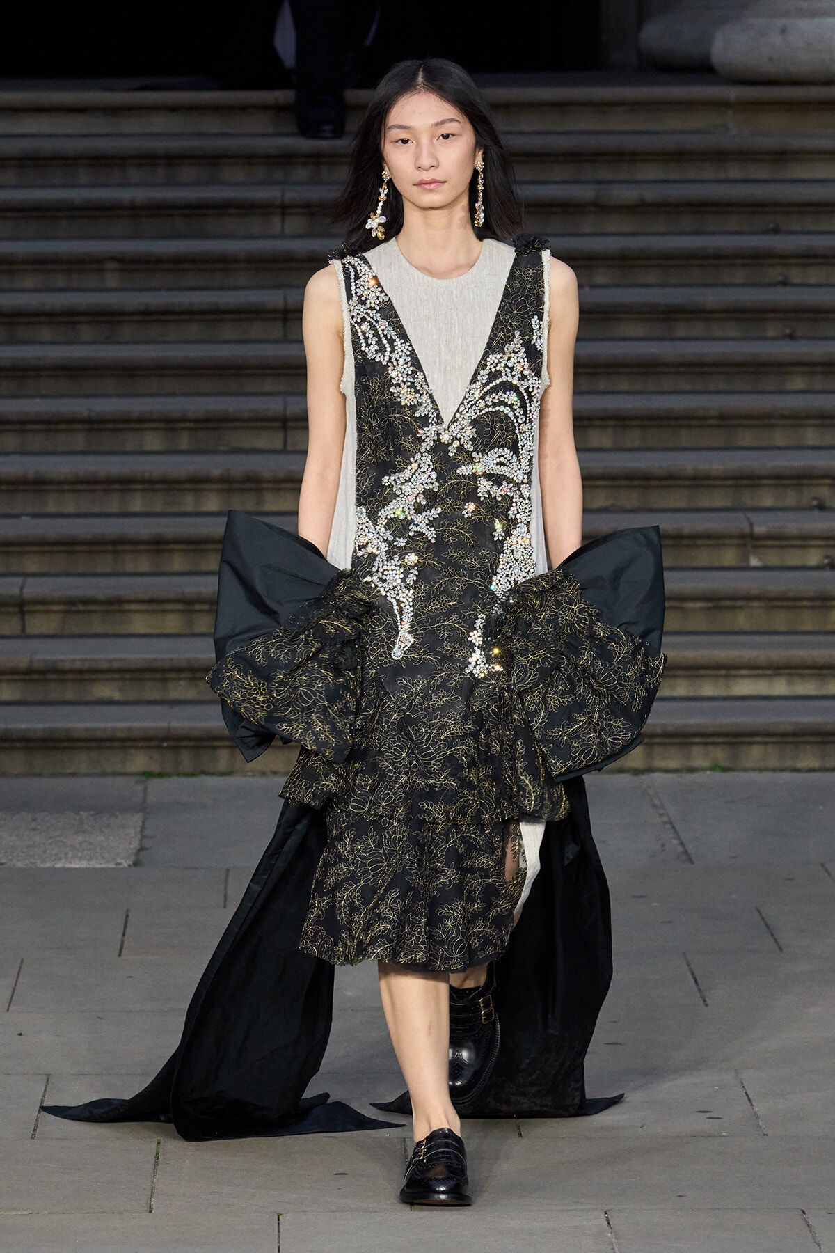 Model walking down a runway in a black-and-gold embroidered gown with a voluminous train and sparkling details, paired with beige inner panel and statement earrings