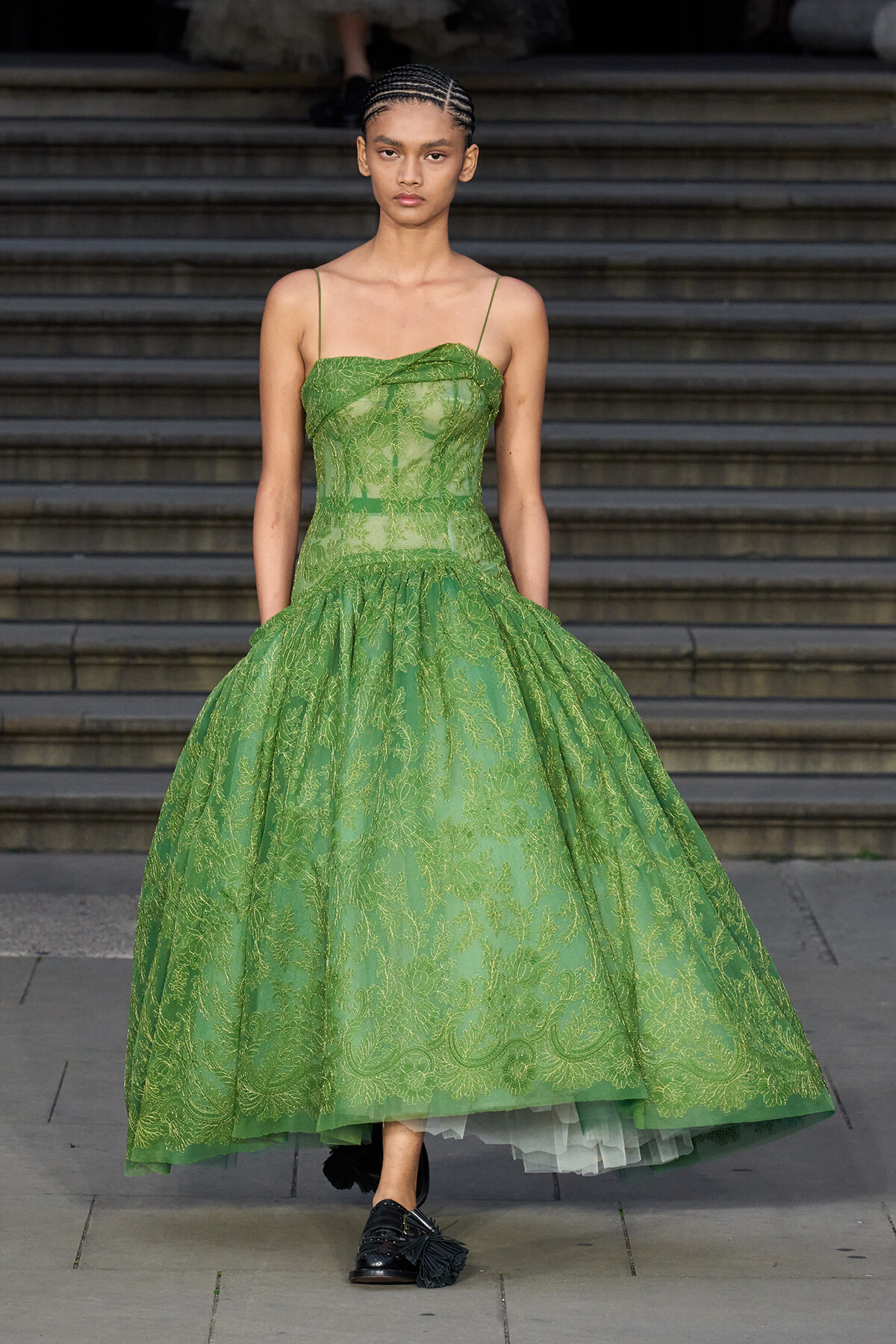 Model walking down stone steps in an emerald-green lace ball gown with a fitted bodice, thin spaghetti straps, and a voluminous skirt.