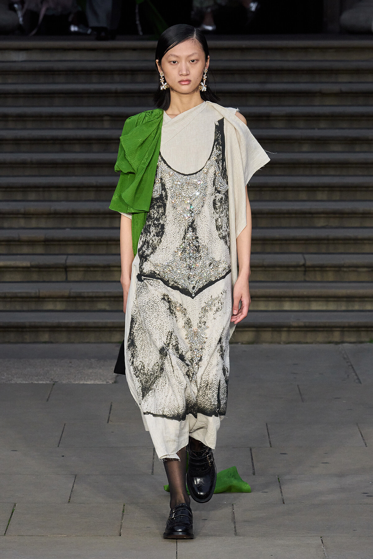 Model on a runway wearing a cream and black beaded dress with a bright green draped sleeve and beige shawl, black loafers, and statement earrings.