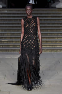 Female model on a runway wearing a sleeveless black crochet dress with long fringe, walking down steps.