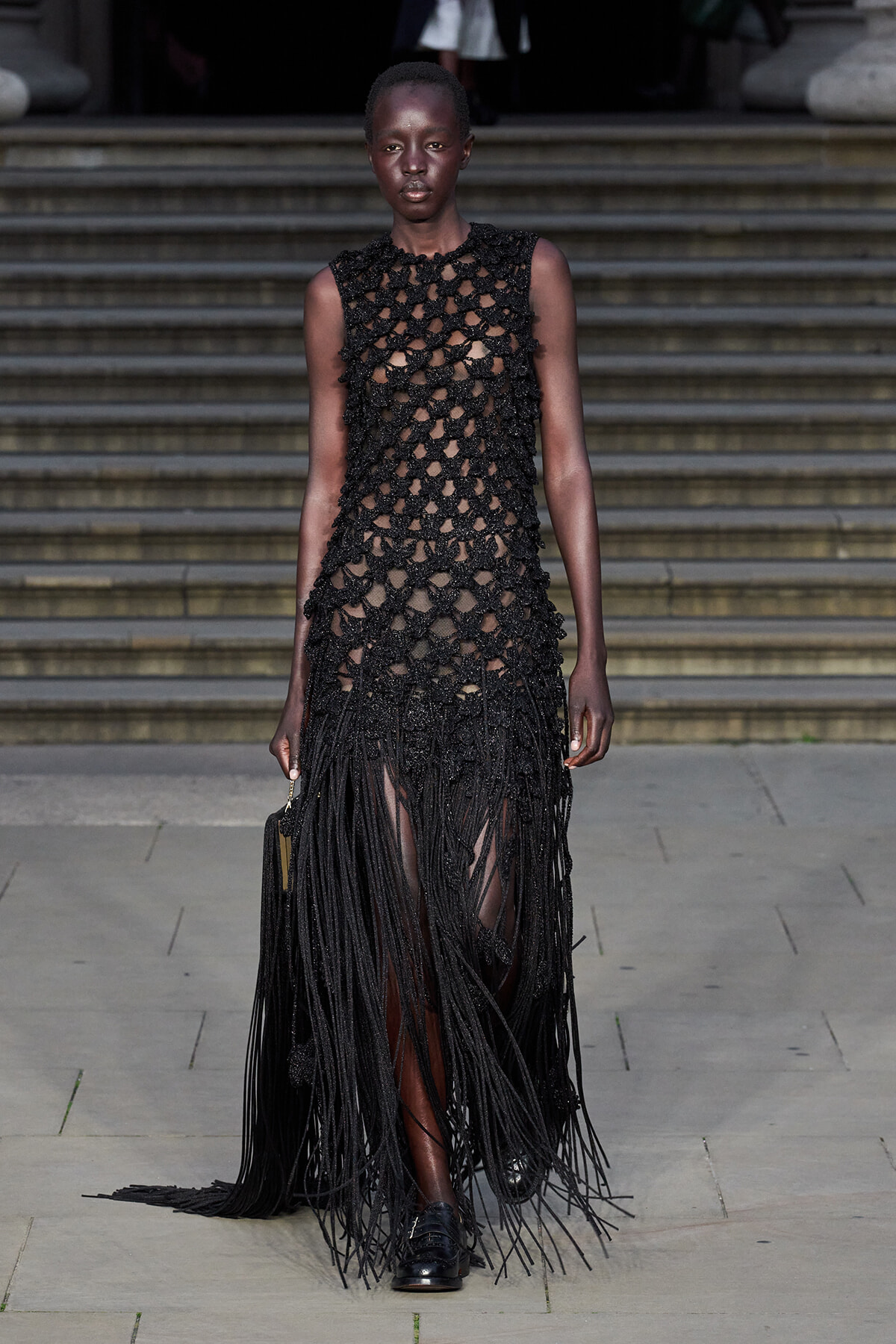 Female model on a runway wearing a sleeveless black crochet dress with long fringe, walking down steps.