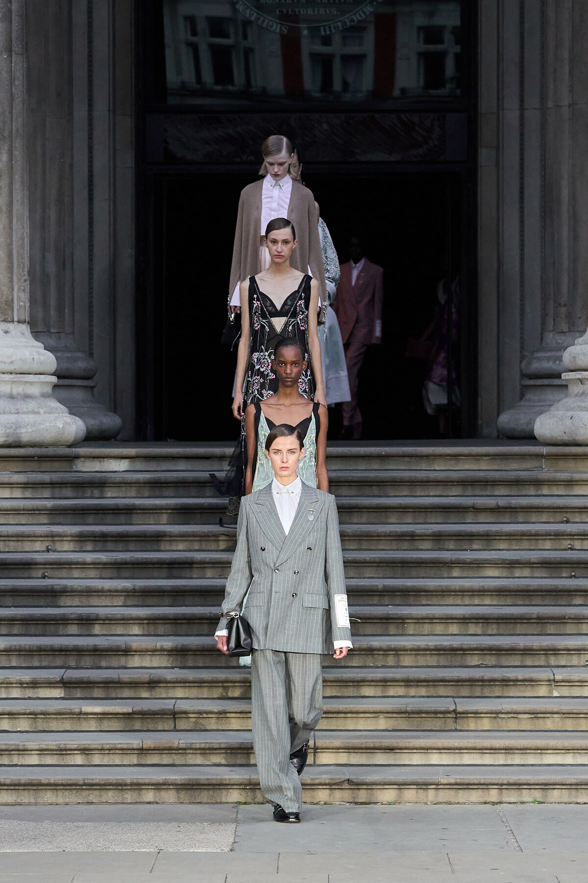 Model in a gray pinstripe suit walks down stone steps at a fashion show, with other models trailing behind on the stairs.