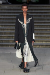 Model on steps wearing an oversized black blazer with jeweled embroidery over a white patterned dress; carries a small black handbag and wears fringed loafers.