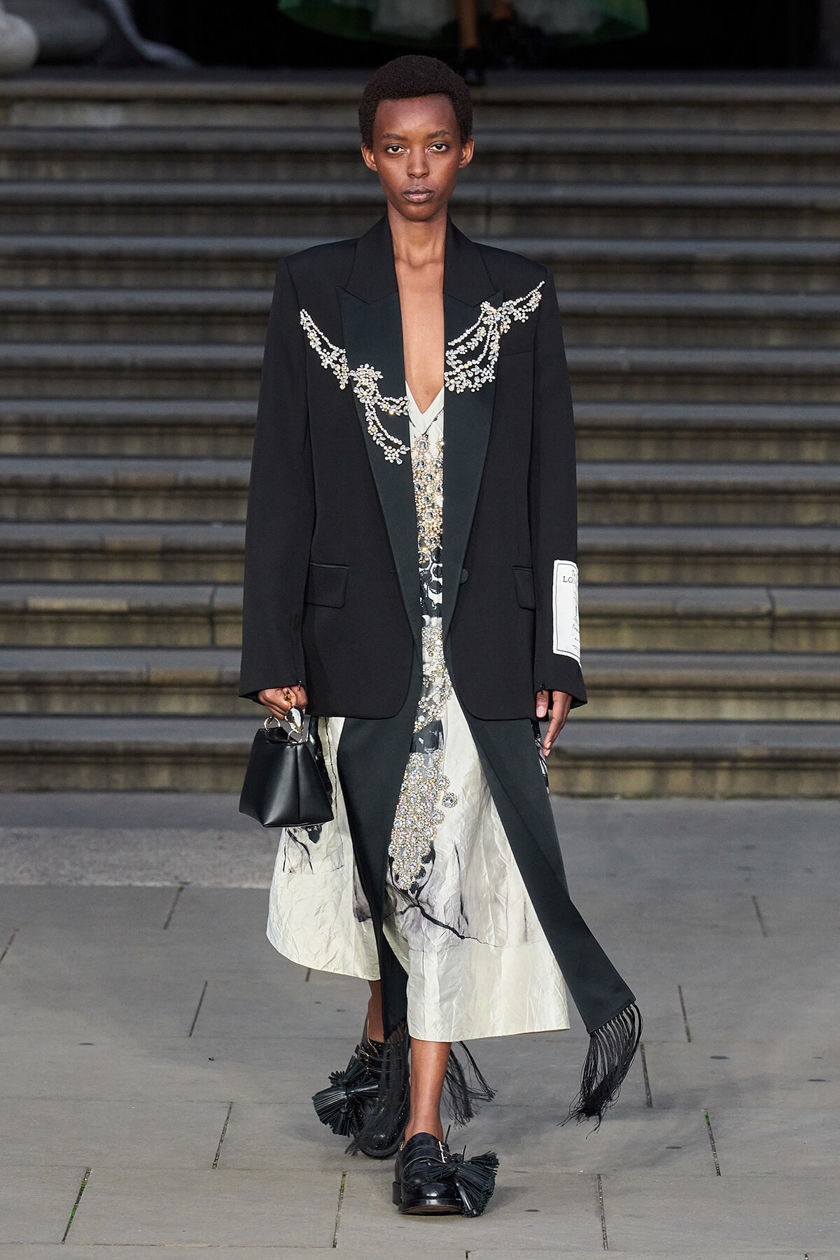 Model on steps wearing an oversized black blazer with jeweled embroidery over a white patterned dress; carries a small black handbag and wears fringed loafers.