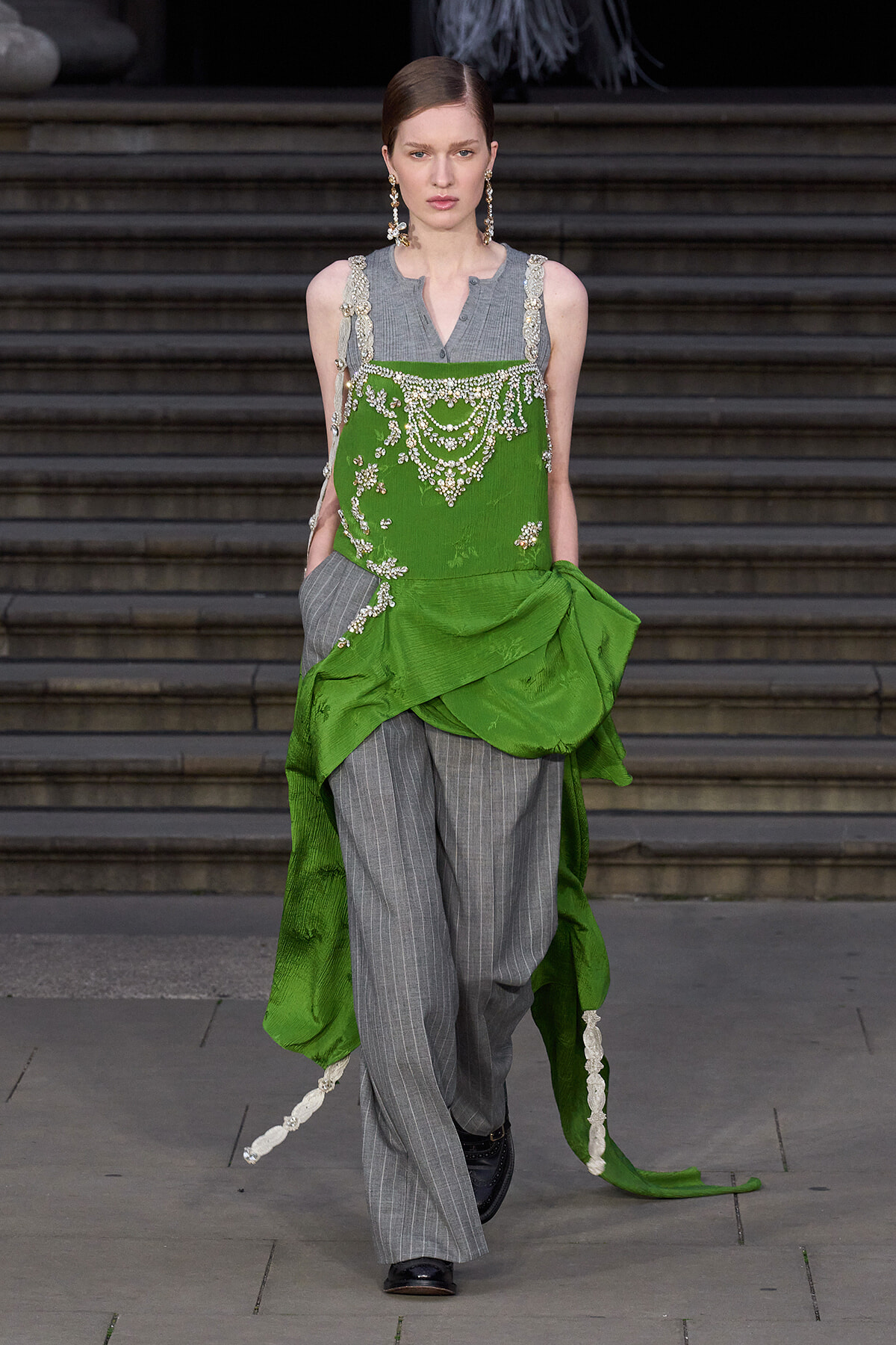Runway model in gray pinstripe trousers with a green embellished overlay and draped sash, featuring pearl embroidery and statement earrings.