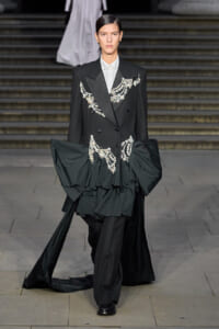 Runway model in a black double-breasted suit with crystal embroidery and a voluminous train at the waist.