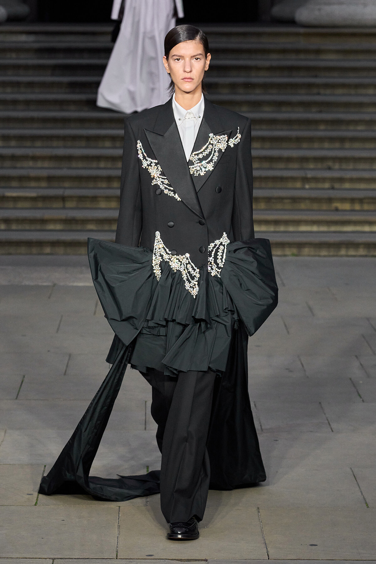 Runway model in a black double-breasted suit with crystal embroidery and a voluminous train at the waist.
