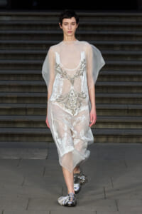 Model walks a runway in a sheer white gown with intricate crystal beadwork and cape-like sleeves, paired with metallic sandals.