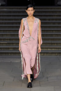 Model walking a pale pink gown with a deep plunging neckline, silver beaded trim, and cape-like sleeves along a stone staircase runway.