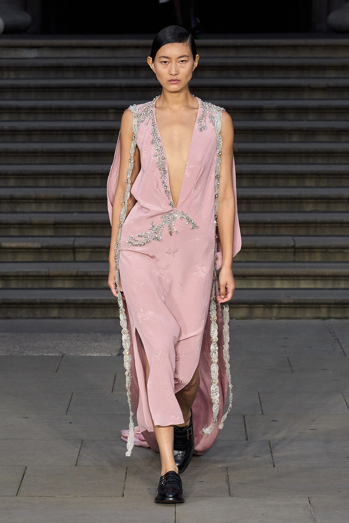 Model walking a pale pink gown with a deep plunging neckline, silver beaded trim, and cape-like sleeves along a stone staircase runway.