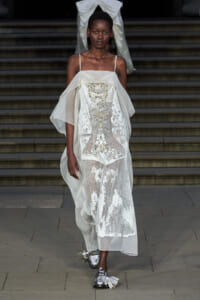 Model walks a fashion runway in a sheer white lace gown with gold embellishments and layered cape-like sleeves, paired with metallic silver tasseled shoes.