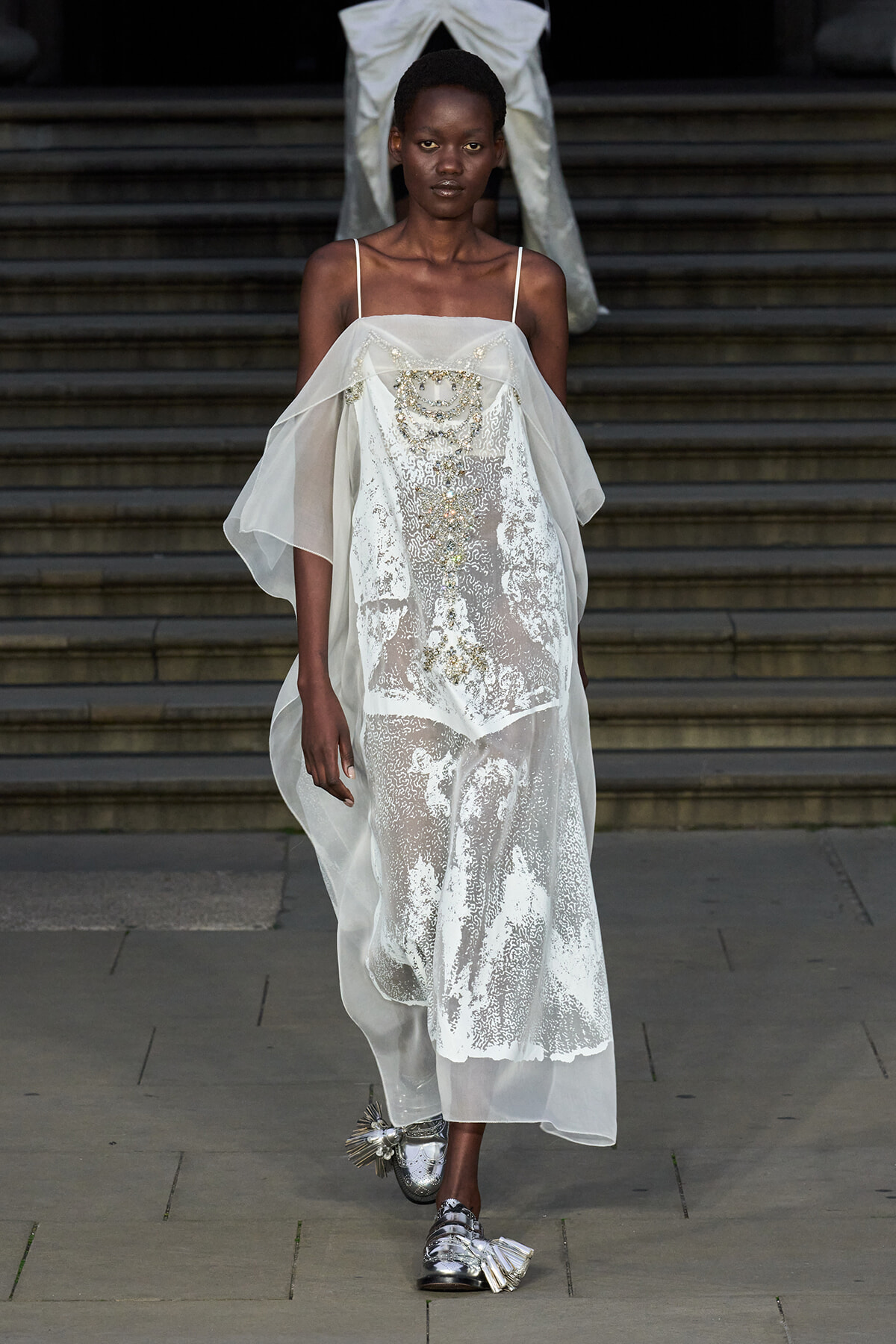 Model walks a fashion runway in a sheer white lace gown with gold embellishments and layered cape-like sleeves, paired with metallic silver tasseled shoes.