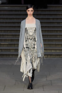 Runway model walks down steps in a light gray cardigan over a cream, beaded dress with silver embroidery and fringe details at the hem.
