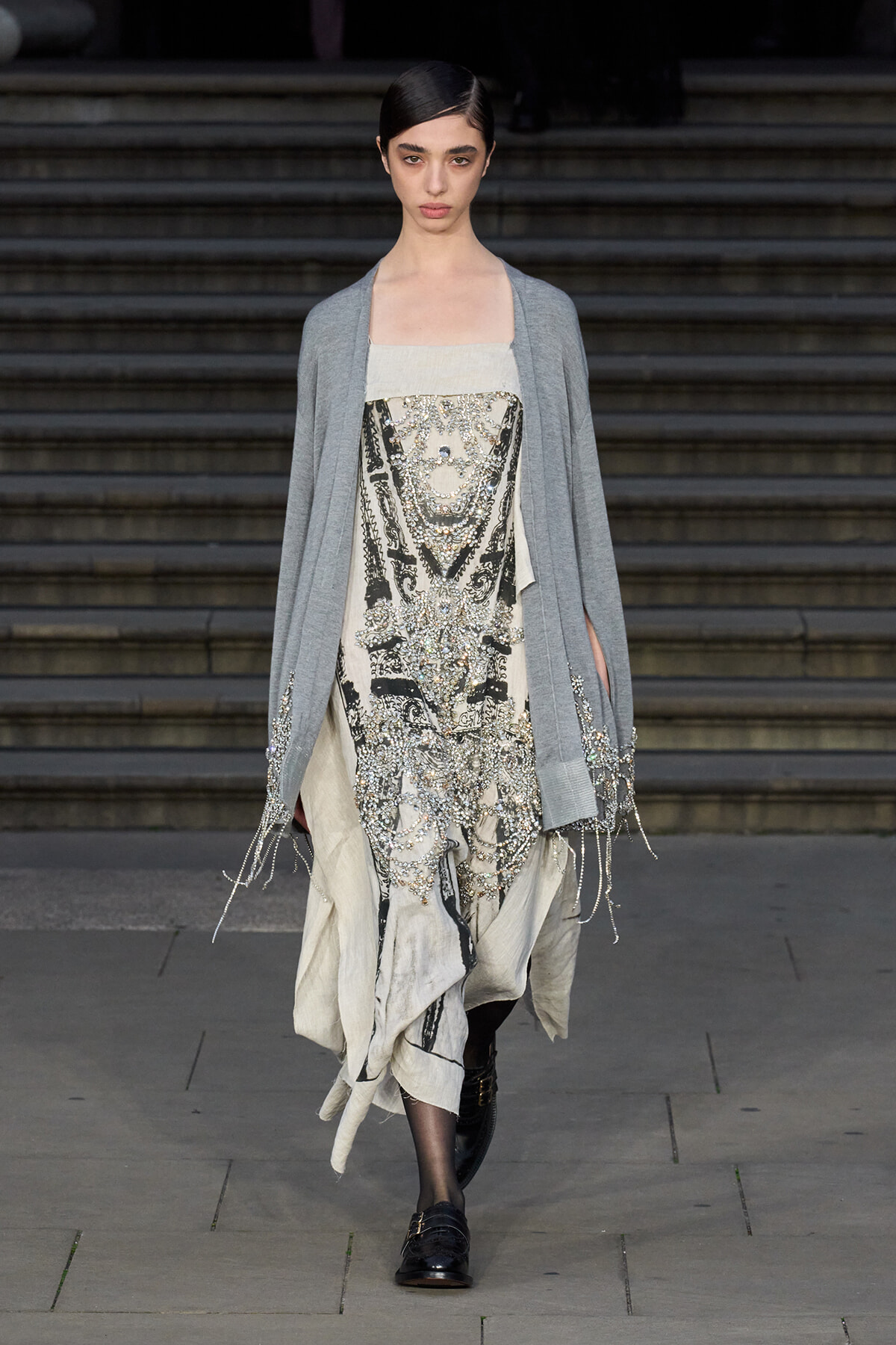 Runway model walks down steps in a light gray cardigan over a cream, beaded dress with silver embroidery and fringe details at the hem.