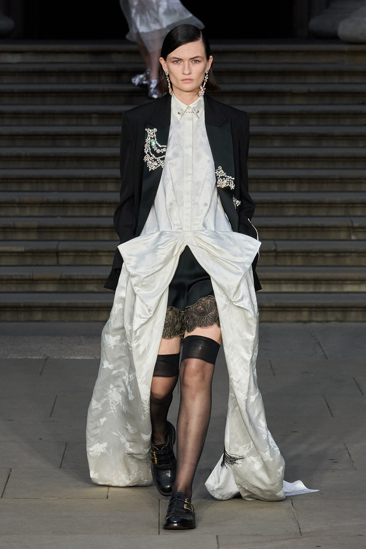 Model walking stairs in a black blazer over a white satin dress with a long, draped train and lace trim, wearing black stockings and loafers with gold buckles.