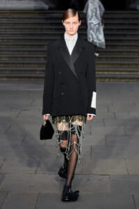 Model walking a runway in a black double-breasted blazer over a white shirt, paired with a sheer beaded chain skirt and black tights, holding a small black handbag.