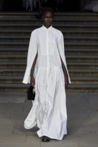 Model in a white floor-length dress with cape-style sleeves walking down a stair-step runway, carrying a small black handbag.