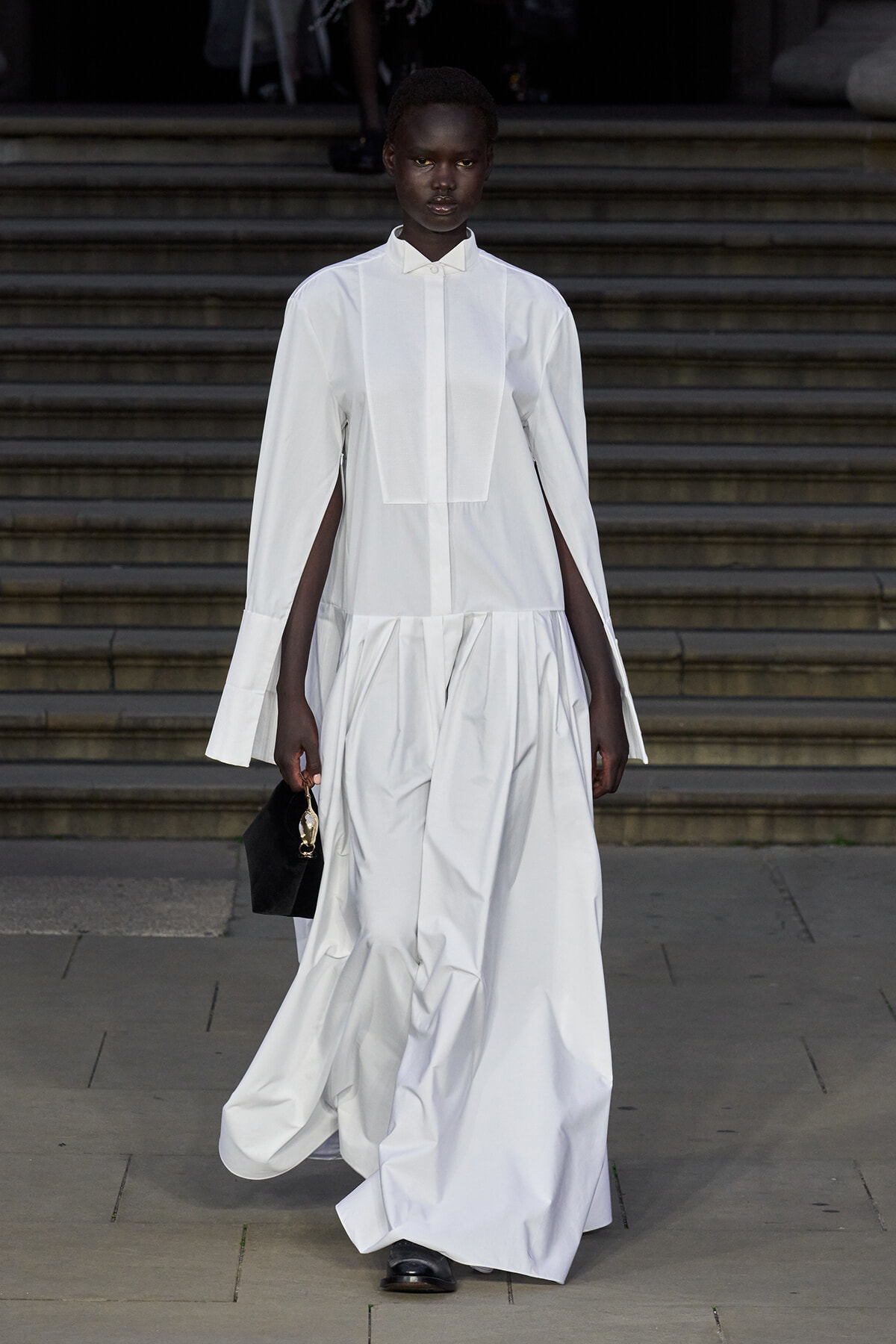 Model in a white floor-length dress with cape-style sleeves walking down a stair-step runway, carrying a small black handbag.