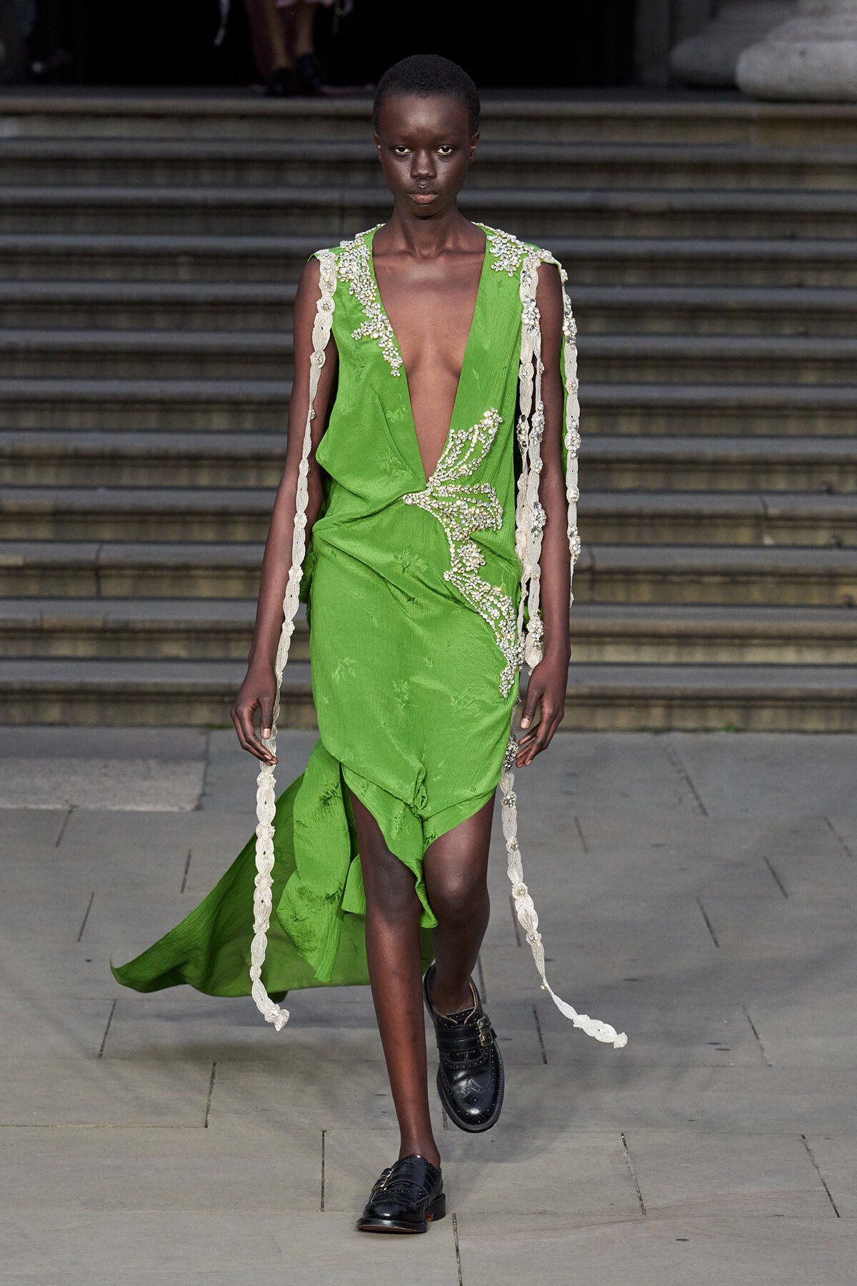 Runway model in a lime-green, deep-V dress with pearl bead embroidery and long cream lace panels trailing at the sides.