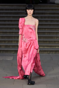 Model on a runway wearing a pink satin one-shoulder gown with floral embroidery and a draped train.