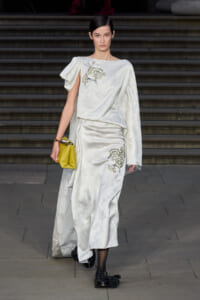 Model walks a runway in a white silk dress with floral embroidery, holding a yellow clutch, descending stone steps in the background.