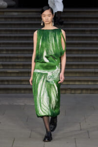 Model walking the runway in a green fringe-dress with a white underlayer, accessorized with large floral earrings.