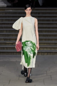 Model on stairs wearing a cream, one-shoulder dress with a bold green floral print at the hem, carrying a pink clutch and wearing black tassel loafers.